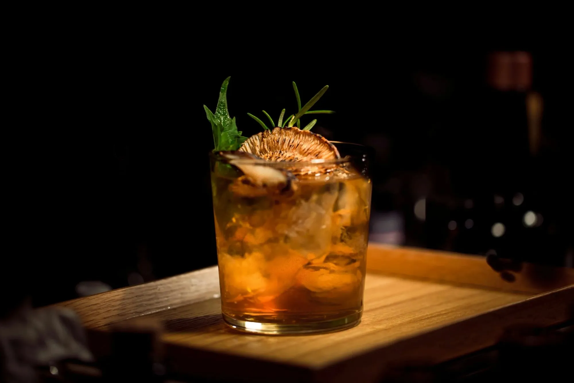 An amber cocktail garnished with dried citrus and herbs on a wooden board.